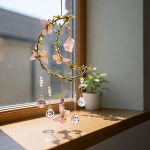 Puede incluir: Un atrapasoles decorativo en forma de media luna. El marco de metal está adornado con flores rosas, hojas verdes y cuentas de cristal colgantes. El atrapasoles está suspendido por una fina cadena dorada.