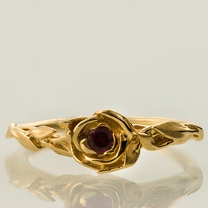 以下が含まれることがあります： 中央に赤い宝石がついた、バラのデザインの金の指輪です。