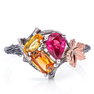 Könnte beinhalten: Silberring mit farbigen Edelsteinen. Das Design umfasst einen tropfenförmigen rosa Stein, zwei rechteckige orangefarbene Steine und einen gelben Stein. Ein roségoldenes Blatt verleiht dem Ring einen natürlichen Look.