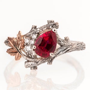 Könnte beinhalten: Ein Ring mit einem birnenförmigen Rubin-Edelstein, der in einer silbernen Fassung mit einem Zweigdesign gefasst ist. Der Ring ist mit einem kleinen Ahornblatt mit einer Roségold-Akzent versehen.