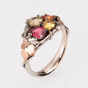 Könnte beinhalten: Ein silberner Ring mit einem filigranen Rankendesign, besetzt mit vier bunten Edelsteinen. Der Ring verfügt über einen roten, gelben und orangefarbenen Edelsteincluster sowie einen einzelnen roten Edelstein.