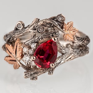 Könnte beinhalten: Ein silberner Ring mit einem roten Edelstein und einem Design aus ineinander verschlungenen Ästen und Blättern. Die Blätter sind aus Gold gefertigt und haben ein detailliertes Design.