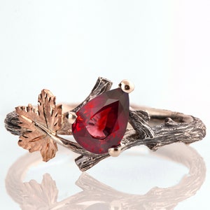 Könnte beinhalten: Silberring mit einem birnenförmigen, tiefroten Edelstein. Der Ring hat ein astähnliches Band mit einem goldfarbenen Blatt und Akzenten. Der Edelstein ist mit kleinen goldenen Zinken gefasst.