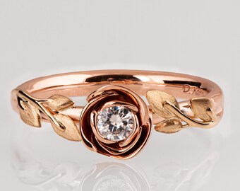 May include: A rose gold ring with a single diamond set in a rose-shaped design. The ring is adorned with delicate leaves on either side of the rose.