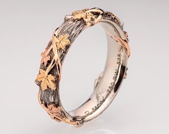 May include: A silver, gold, and rose gold band ring with a textured design of a tree branch with leaves. The inside of the band is stamped "D. Kerav - 750 SW - 585".