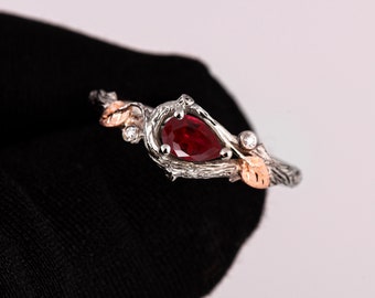 May include: A pear-shaped ruby gemstone set in a silver ring with a twig-like band and two small diamonds. The ring is accented with two gold leaves.