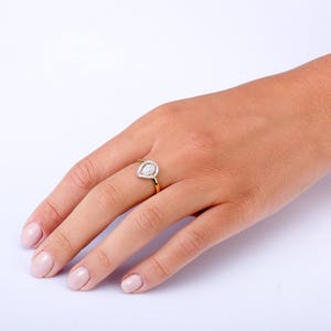 Puede incluir: Un anillo de oro con una piedra clara en forma de pera engastada en un halo de piedras claras m&aacute;s peque&ntilde;as.
