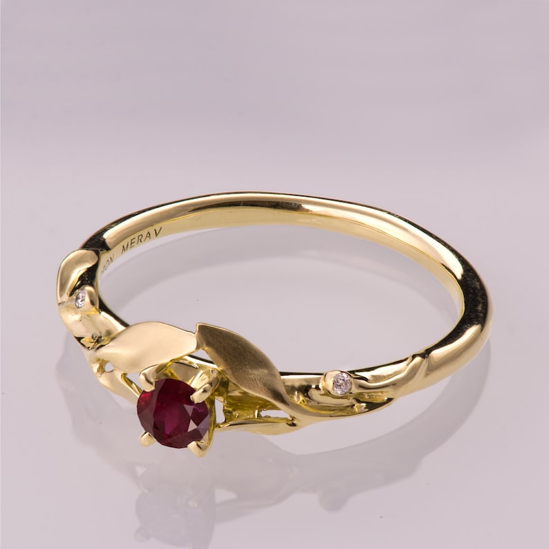 May include: A gold ring featuring a ruby gemstone and two small diamonds set in a leaf design. The ring is engraved with the word "MERAV".