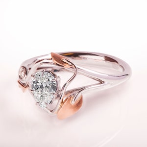 May include: A pear-shaped diamond engagement ring set in a white gold band with rose gold accents. The ring features a delicate floral design with leaves and a small diamond accent.