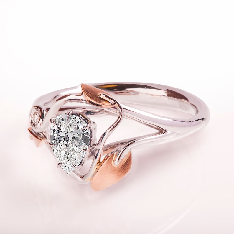 May include: A pear-shaped diamond engagement ring set in a white gold band with rose gold accents. The ring features a delicate floral design with leaves and a small diamond accent.