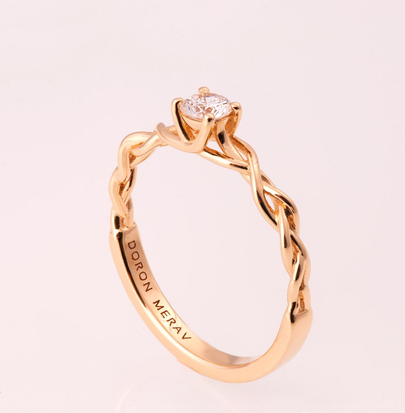 K&ouml;nnte beinhalten: Ein goldener geflochtener Ring mit einem einzelnen runden Diamanten, der in der Mitte sitzt. Der Ring ist auf der Innenseite mit den Worten "DORON MERAV" graviert.