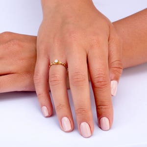 K&ouml;nnte beinhalten: Ein goldener Ring mit einem einzigen, kleinen, runden Diamanten, der in der Mitte sitzt. Der Ring befindet sich an einem Finger einer Person, und die N&auml;gel der Person sind in einem hellen Rosaton lackiert.