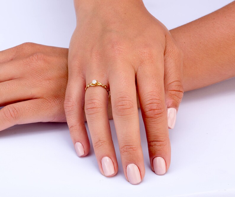 K&ouml;nnte beinhalten: Ein goldener Ring mit einem einzigen, kleinen, runden Diamanten, der in der Mitte sitzt. Der Ring befindet sich an einem Finger einer Person, und die N&auml;gel der Person sind in einem hellen Rosaton lackiert.