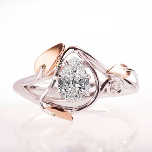 May include: A pear-shaped diamond engagement ring set in a white gold band with a rose gold leaf design. The ring features a delicate, intricate design with a single diamond accent.