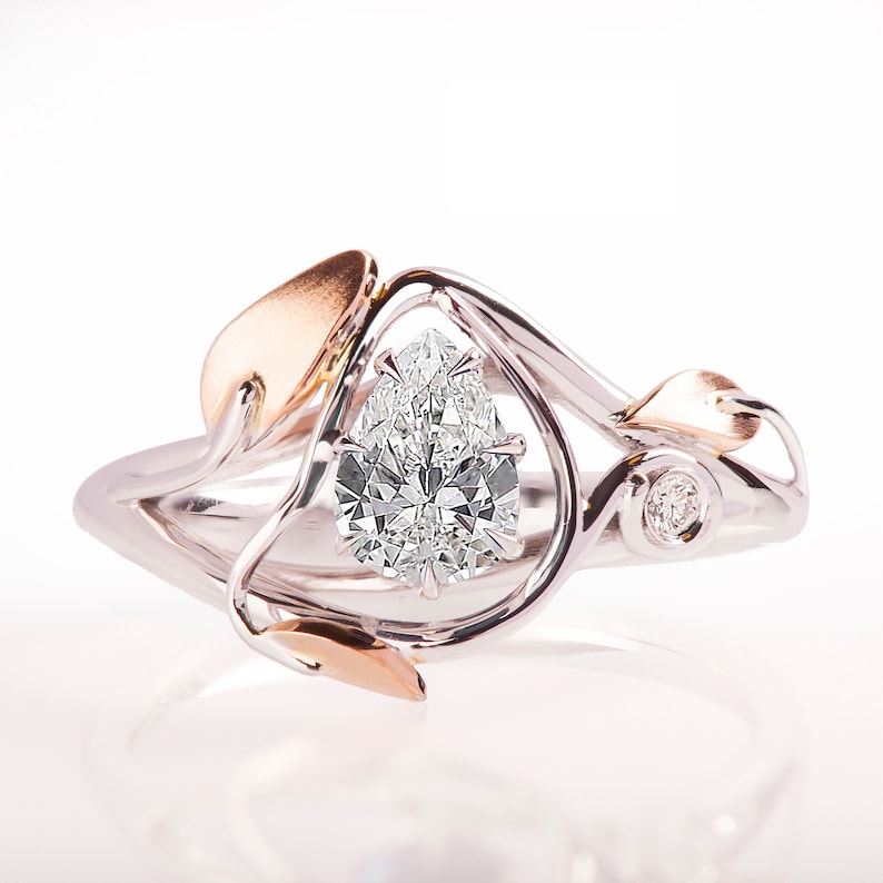 May include: A pear-shaped diamond engagement ring set in a white gold band with a rose gold leaf design. The ring features a delicate, intricate design with a single diamond accent.