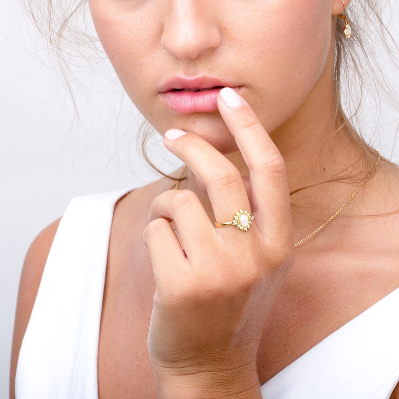 May include: A gold ring with a large white pearl surrounded by smaller white pearls and gold accents. The ring is on a person's finger.