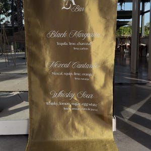 May include: A gold-colored bar menu with the text "A Bar" at the top. The menu lists three cocktails: Black Margarita, Mezcal Cantarito, and Whisky Sour, with their ingredients. The background is a gold fabric.