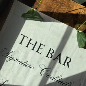 May include: Off-white canvas bar sign with the words "THE BAR Signature Cocktails" in black script. The sign is partially lit by sunlight, with a wooden surface in the background. Green leaves are visible.