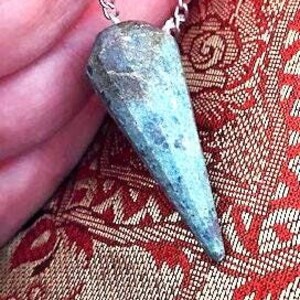 Könnte beinhalten: Eine silberne Kette mit einem blau-grünen Kristall-Anhänger in Form einer Spitze. Der Anhänger ist kegelförmig und hat eine raue, strukturierte Oberfläche.