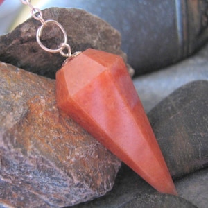 May include: A red carnelian crystal pendulum on a silver chain. The pendulum is faceted and has a pointed tip. It is resting on a rock.