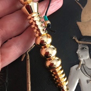 Könnte beinhalten: Ein goldfarbenes Metallpendel mit einer spitzen Spitze und einer Reihe runder Perlen. Das Pendel ist an einer schwarzen Schnur mit einer türkisfarbenen Perle befestigt.