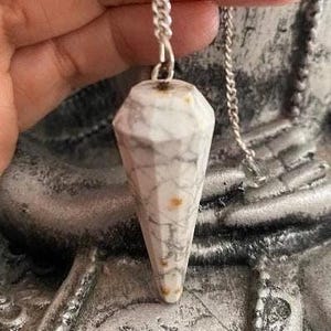 May include: White marble pendulum with a silver chain. The pendulum is shaped like a pointed cone with a faceted design.