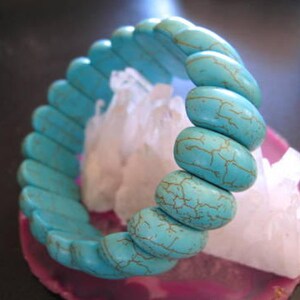 May include: Turquoise blue bracelet made of smooth, oval-shaped beads with a mottled, marbled pattern. The bracelet is stacked on a white crystal.