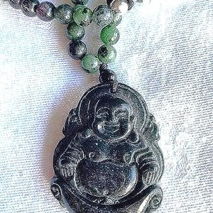 May include: A dark, polished stone pendant carved into the shape of a smiling Buddha figure, suspended from a beaded necklace featuring round, mottled green and dark purple stone beads with a smooth, glossy finish and a knotted cord attachment.