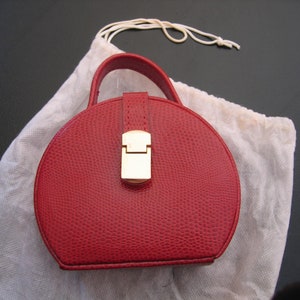 May include: A red, round handbag with a textured surface and a gold-colored clasp. The bag has a small handle and is partially inside a white drawstring bag. The bag is a vibrant red color.