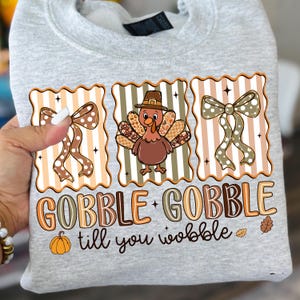 May include: A light gray sweatshirt featuring a Thanksgiving-themed design. The design includes a cartoon turkey wearing a pilgrim hat, two bows, and the text "GOBBLE GOBBLE till you wobble" in brown and orange.