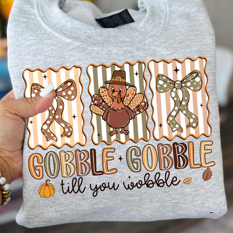 May include: A light gray sweatshirt featuring a Thanksgiving-themed design. The design includes a cartoon turkey wearing a pilgrim hat, two bows, and the text "GOBBLE GOBBLE till you wobble" in brown and orange.