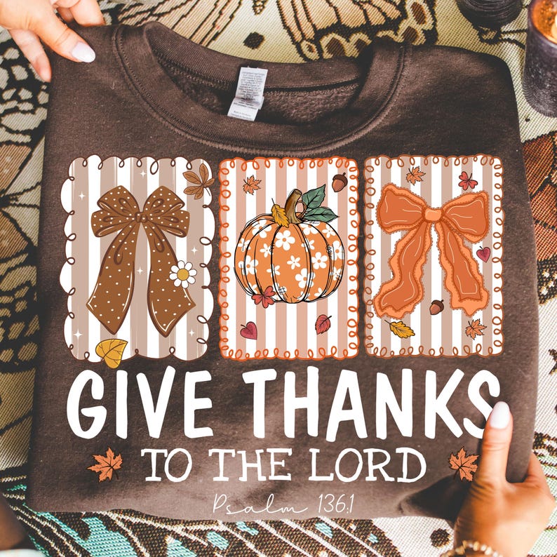Pode incluir: Um moletom marrom com um design tem&aacute;tico de outono. O design inclui tr&ecirc;s pain&eacute;is retangulares com um la&ccedil;o, uma ab&oacute;bora e outro la&ccedil;o. O texto "GIVE THANKS TO THE LORD" &eacute; impresso abaixo, com "Psalm 136:1".