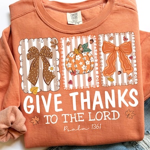 Pode incluir: Camisola laranja com um design gr&aacute;fico com la&ccedil;os, uma ab&oacute;bora e folhas de outono. O texto "GIVE THANKS TO THE LORD" &eacute; impresso em branco, com "Psalm 136:1" abaixo. Uma etiqueta Comfort Colors &eacute; vis&iacute;vel.