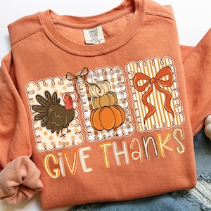 May include: Orange sweatshirt with a Thanksgiving-themed graphic. The graphic includes a turkey, stacked pumpkins, and a bow, all within decorative frames. The words "GIVE THANKS" are printed in a colorful font.