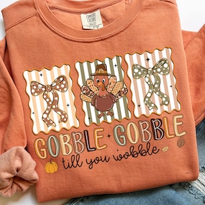 May include: Orange Comfort Colors sweatshirt with a Thanksgiving-themed graphic. The design features a turkey wearing a pilgrim hat, bows, and the text "GOBBLE GOBBLE till you wobble" in a playful font. A pumpkin and leaves are also included.