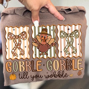 May include: A brown long-sleeve shirt with a Thanksgiving-themed graphic. The graphic features a turkey wearing a hat, bows, a pumpkin, and the text "GOBBLE GOBBLE till you wobble". The design is in shades of brown, orange, and white.