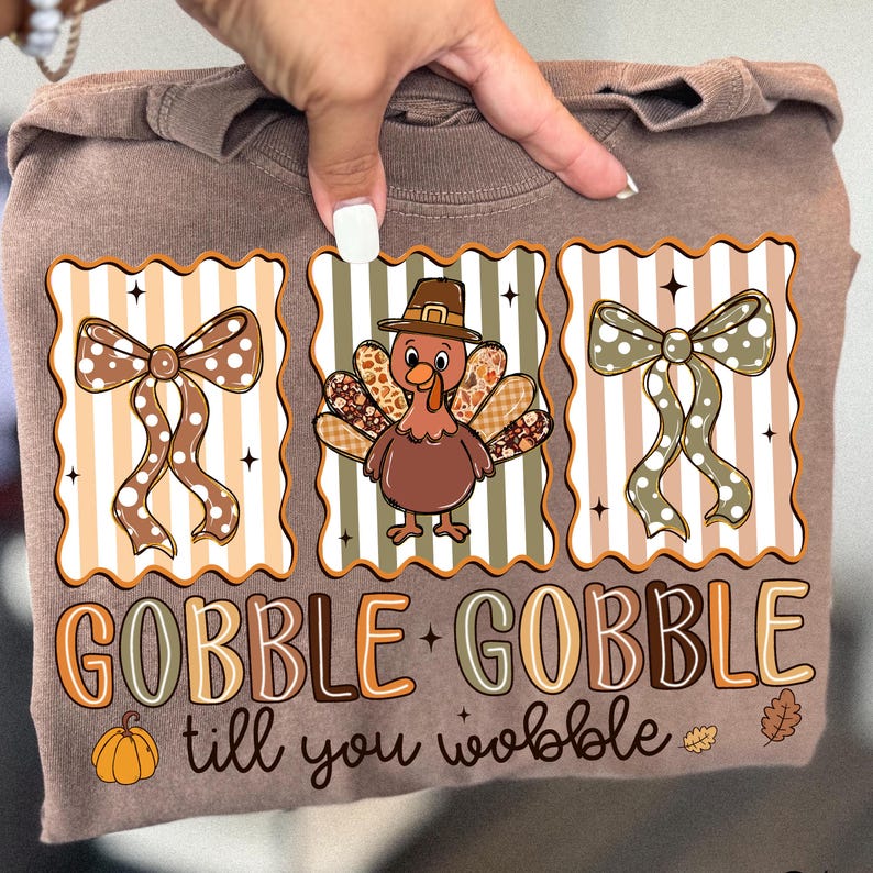 May include: A brown long-sleeve shirt with a Thanksgiving-themed graphic. The graphic features a turkey wearing a hat, bows, a pumpkin, and the text "GOBBLE GOBBLE till you wobble". The design is in shades of brown, orange, and white.