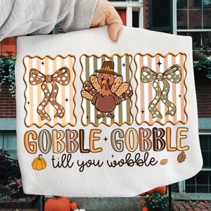 May include: White Thanksgiving-themed towel with a turkey wearing a pilgrim hat, two decorative bows, and the text "GOBBLE + GOBBLE till you wobble". The design features orange, brown, and beige colors.