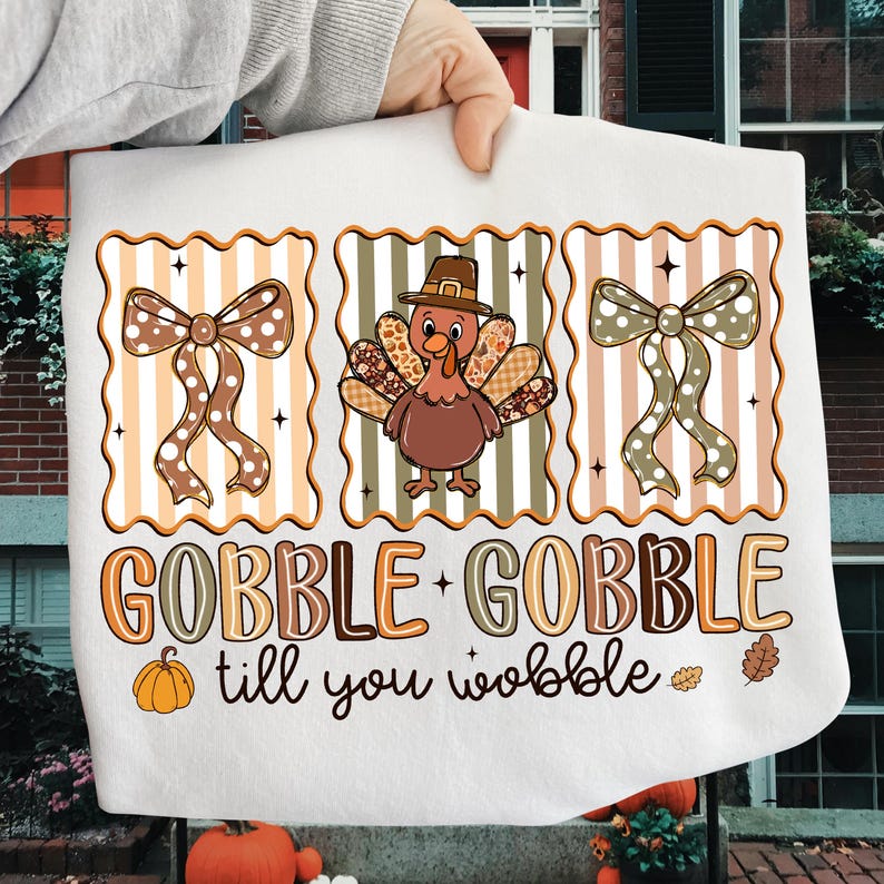 May include: White Thanksgiving-themed towel with a turkey wearing a pilgrim hat, two decorative bows, and the text "GOBBLE + GOBBLE till you wobble". The design features orange, brown, and beige colors.