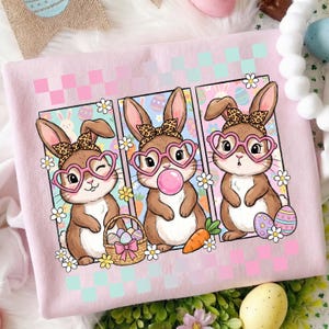 May include: Pink sweatshirt featuring three cartoon bunnies wearing heart-shaped glasses and leopard print bows. Each bunny is framed with flowers and a checkered background. One bunny has a basket of Easter eggs.