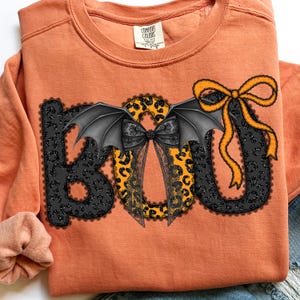 May include: Orange sweatshirt with a Halloween-themed "BOO" design. The letters are black with a lace pattern, leopard print, and bat wings. An orange bow is on the right side of the "U". The sweatshirt has a crew neck and long sleeves.