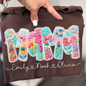 Könnte beinhalten: Ein braunes Sweatshirt mit dem Wort "MOM" in bunten Buchstaben mit Blumenmuster. Die Buchstaben sind mit weißer Spitze umrandet und mit rosa Schleifen verziert. Unter dem Wort "MOM" stehen die Namen "Emily", "Noah" und "Olivia" in Schreibschrift.