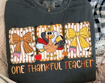 Könnte beinhalten: Dunkelgraues Sweatshirt mit einer farbenfrohen Grafik mit einer Truthahn, Bleistiften und Schleifen. Der Text "ONE THANKFUL TEACHER" ist in orangefarbenen und braunen Buchstaben dargestellt. Das Design befindet sich auf einem gestreiften Hintergrund.