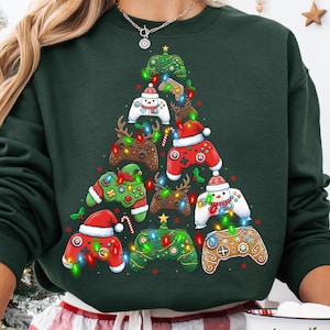 May include: Dark green sweatshirt with a Christmas tree design composed of video game controllers. The controllers are adorned with festive decorations such as Santa hats, lights, and reindeer antlers. A fun holiday themed apparel item.