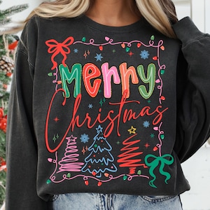 May include: Dark gray sweatshirt with a colorful "Merry Christmas" graphic. The design includes Christmas trees, string lights, snowflakes, and bows in red, green, pink, and blue. A festive holiday-themed apparel item.