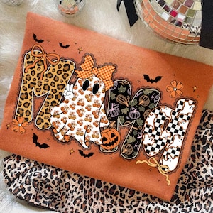 May include: An orange Halloween sweatshirt with the word "MAMa" in various patterns. The letters include leopard print, pumpkin patterns, and a checkered design. Black bats and orange flowers decorate the shirt.