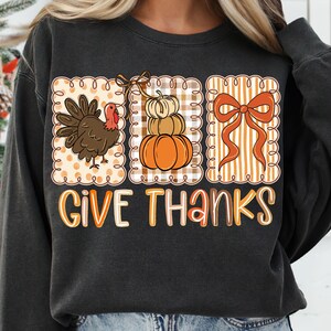 May include: Dark gray sweatshirt with a Thanksgiving-themed graphic. The graphic features a turkey, a stack of pumpkins, and a bow, all within decorative frames. The words "GIVE THANKS" are written in orange and white letters.