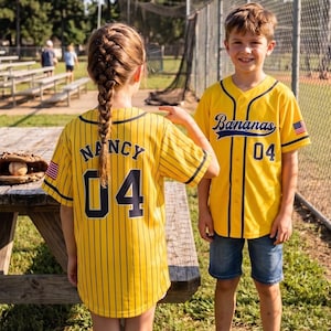 Könnte beinhalten: Zwei Kinder tragen gelbe Baseball-Trikots mit schwarzen Nadelstreifen und der Nummer 04. Auf einem Trikot steht "Nancy" auf dem Rücken, auf dem anderen "Bananas". Beide haben einen amerikanischen Flaggenaufnäher auf dem Ärmel. Ein Baseballhandschuh und ein Ball liegen auf einem Picknicktisch aus Holz.