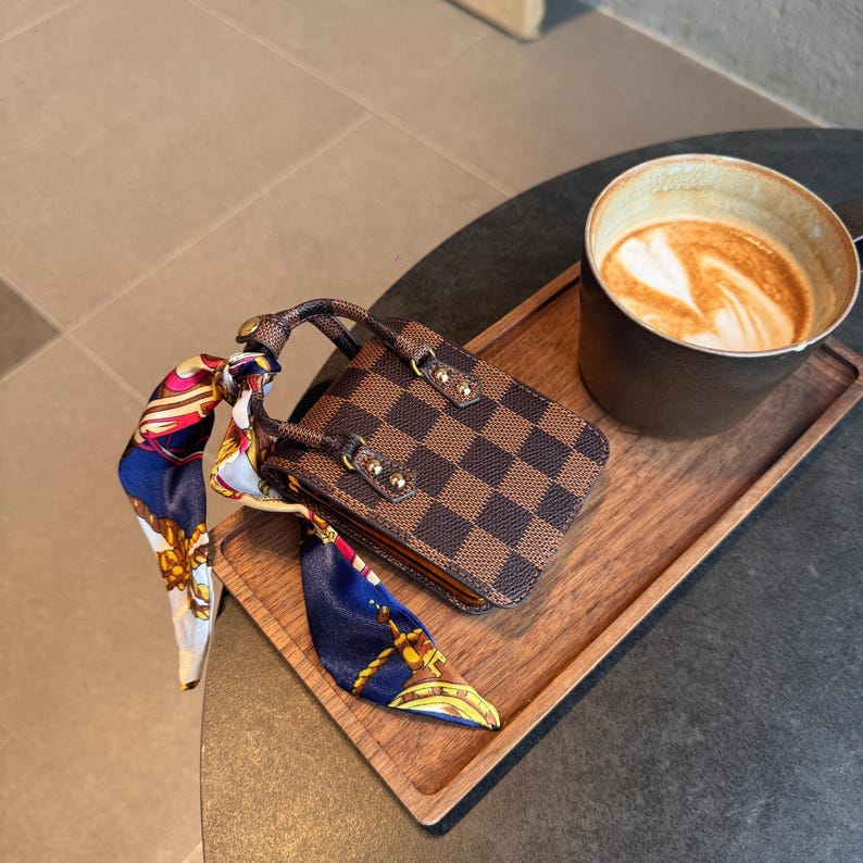 May include: A miniature brown and tan checkered handbag with a decorative scarf, resting on a wooden tray next to a cup of coffee. The handbag has a handle and gold-colored hardware. The scarf is blue, red, and gold.