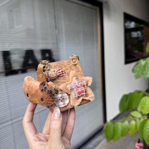 May include: A small, brown handbag with a gold chain strap, adorned with a plush Hello Kitty, a star-shaped patch with "Sweet Cherry" text, and a button. The bag has a textured surface and a small handle.